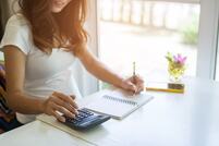 Femme utilisant une calculatrice et prenant des notes à une table blanche près d'une fenêtre ensoleillée avec une petite plante en pot.